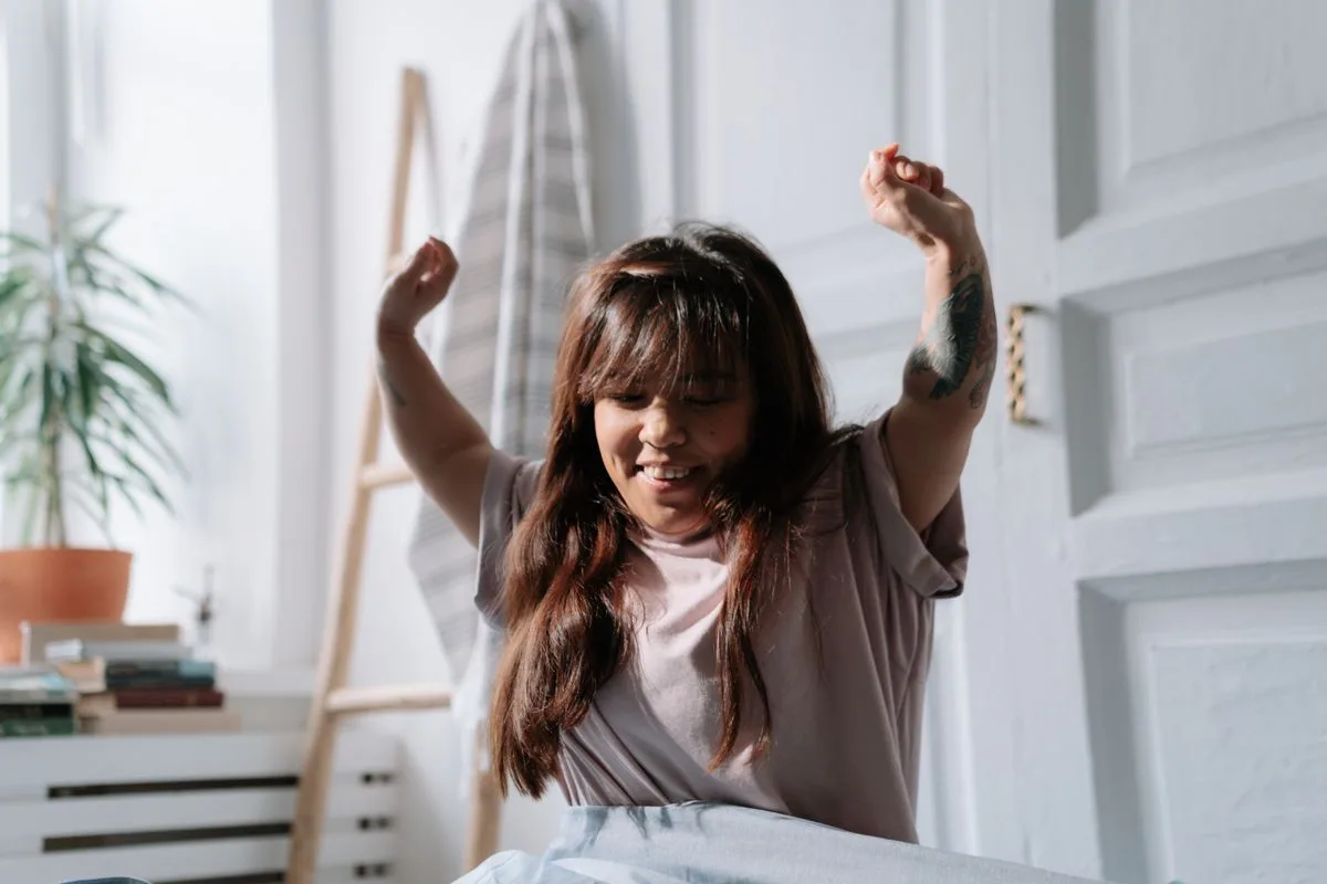 Relaxed Asian woman wakes up and stretches in a sunlit bedroom, enjoying the morning light