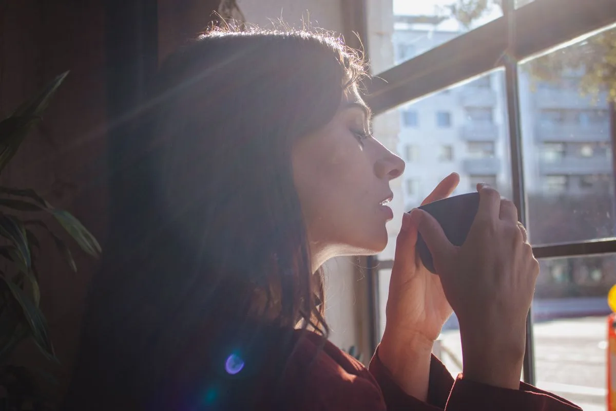 Woman savoring a hot drink by a sunlit window in the morning, calm urban setting